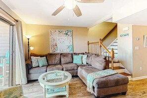 Ein Wohnzimmer mit Ecksofa, einem Glastisch, einer Treppe mit Holzbalken und einem Deckenventilator.