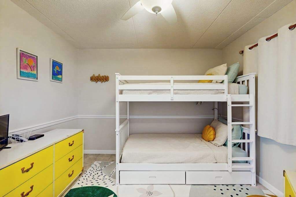 Une chambre avec un lit superposé, un bureau et des tiroirs, un ventilateur de plafond et deux tableaux accrochés au mur.