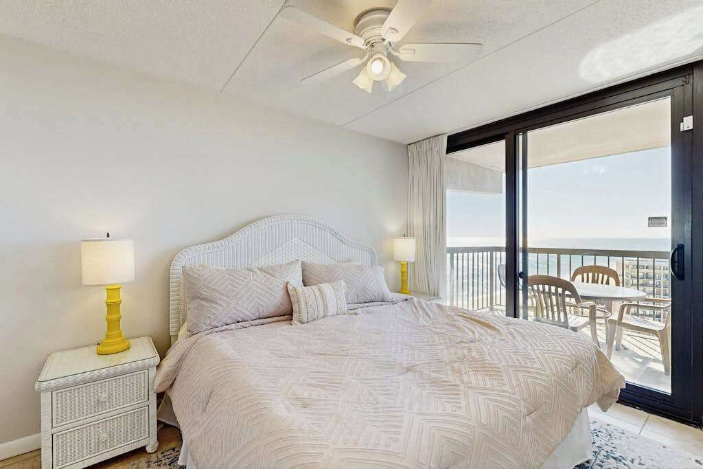 A bedroom with a bed, two bedside lamps, a ceiling fan, and a balcony with chairs and a table.