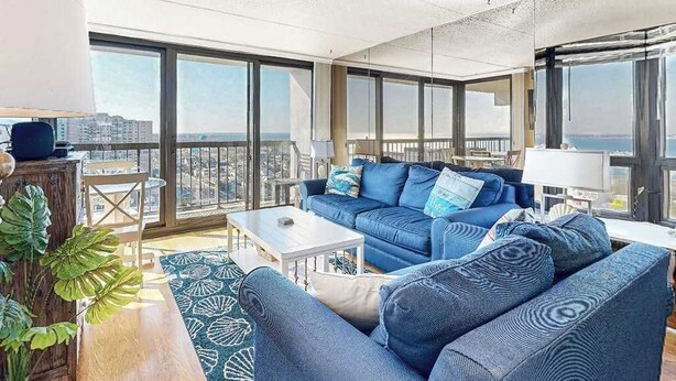 A modern living room with a blue sofa, a coffee table, and a view of the city through large windows. - Coastal-themed beach condo with balcony, views, pools, tennis & central AC (Ocean City)