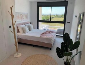 Ein Schlafzimmer mit einem Bett, einem großen Fenster, einer Pflanze und einer Stehlampe.