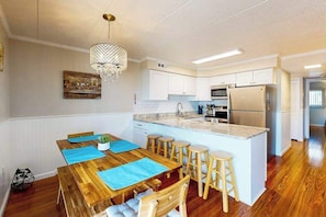 A modern kitchen with white cabinets, a central island with stools, a chandelier, and a dining table with blue placemats. - Salty Sands II 103 (Ocean City)