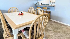 Un comedor con una mesa blanca, sillas de madera, piso alfombrado y una zona de cocina con microondas y utensilios.