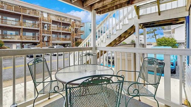En balkon med et glasbord og metalstole, der kigger ud over en gade med bygninger og parkerede biler.