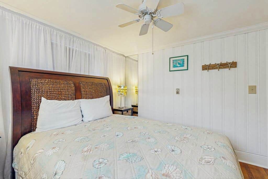 A bedroom with a bed, a ceiling fan, a nightstand, a lamp, a framed picture, and a wall-mounted hook.