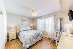 Une chambre à coucher comprenant un lit, des tables de chevet, une chaise, un ventilateur de plafond et une fenêtre avec des rideaux.