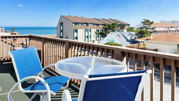 En balkon med to stole og et bord, med udsigt over en strand og bygninger.