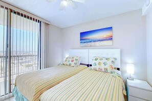 Une chambre d’hôtel avec un grand lit, un ventilateur de plafond, un tableau représentant un coucher de soleil et une vue par la fenêtre.