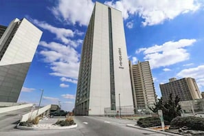A modern building with a prominent vertical design, a clear sky, and a parking area with a few vehicles. - 16th-floor ocean-view condo with game room, in/outdoor pools, tennis, saunas, AC (Ocean City)