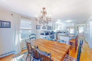 Un comedor con una mesa y sillas de madera, una araña y una cocina al fondo.