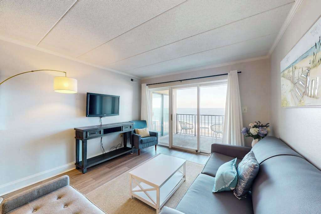 A modern living room with a flat-screen TV, a sofa, a coffee table, and a balcony with a view.