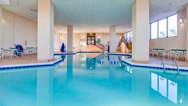 Una piscina interna con acqua cristallina, circondata da pareti e colonne bianche, con una scala che conduce a un livello superiore.