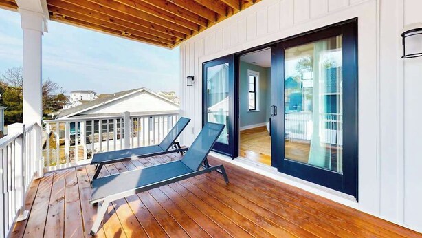 Une terrasse en bois avec deux chaises longues, une porte vitrée donnant sur une pièce et un mur extérieur blanc.