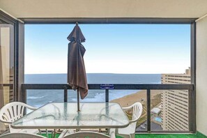 A balcony with a glass table and chairs, a brown umbrella, and a view of the ocean and buildings. - Oceanfront escape with beach access, pools, sauna, tennis, & fitness room (Ocean City)