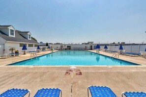 En rektangulär swimmingpool med blå solstolar och parasoller på båda sidor, omgiven av en betongplattform.