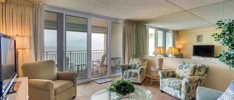 Quarto de hotel com varanda, sofá, poltrona, mesa de centro e televisão.