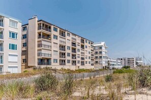 Un edificio residencial de varios pisos con balcones, una playa arenosa en primer plano y cielos despejados.