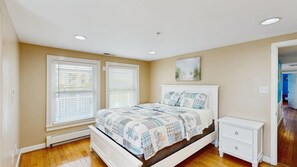 A bedroom with a bed, two windows with blinds, a nightstand, and a painting on the wall. - Boardwalk Bungalow (Ocean City)