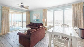 Una habitación con sofá, una mesa de comedor con sillas, ventilador de techo y ventanas amplias con cortinas.