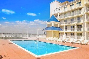 A swimming pool with lounge chairs, a building with balconies, and a clear sky. - Belmont Towers 601 (Ocean City)