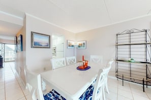 A dining area with a white table, white chairs, and a black metal shelf unit. - Sixth-floor oceanfront condo w/central AC, washer/dryer, furnished balcony (Ocean City)