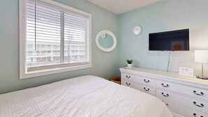 A bedroom with a bed, a dresser, a round mirror, a wall-mounted TV, and a window with blinds. - Jocelyn Manor Unit 85 (Ocean City)