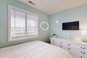Ein Schlafzimmer mit einem Bett, einem Schminktisch, einem runden Spiegel, einem an der Wand befestigten Fernseher und einem Fenster mit Jalousien.