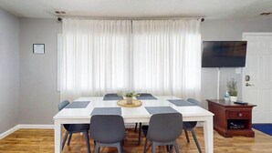 A room with a white table, four chairs, a flat-screen TV, a wooden cabinet, and a window with white curtains. - Jocelyn Manor Unit 85 (Ocean City)