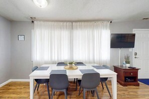 Ein Zimmer mit einem weißen Tisch, vier Stühlen, einem Flachbildfernseher, einem Holzschrank und einem Fenster mit weißen Vorhängen.