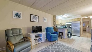A living room with a sofa, TV, and blue armchair, a kitchen with cabinets and a refrigerator, and artwork on the walls. - Constellation House 107 (Ocean City)