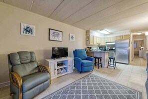 Ein Wohnzimmer mit Sofa, Fernseher und blauem Sessel, eine Küche mit Schränken und Kühlschrank, sowie Kunstwerken an den Wänden.