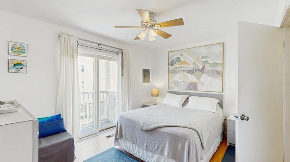 A hotel room with a bed, a desk, a chair, a ceiling fan, a window with curtains, and a painting on the wall. - Alexandra 103 (Ocean City)