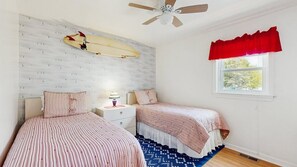 A bedroom with two beds, a ceiling fan, a surfboard on the wall, and a window with a red valance. - Alexandra 103 (Ocean City)