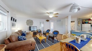 A living room with a sofa, chairs, a coffee table, and a dining table with chairs. There is a ceiling fan, a television, and a kitchen area in the background. - Alexandra 103 (Ocean City)