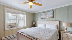 A bedroom with a wooden bed, a ceiling fan, a window with blinds, a nightstand with a lamp, and a wall decoration. - Homey, comfortable condo w/ a furnished balcony - just blocks from the beach (Ocean City)
