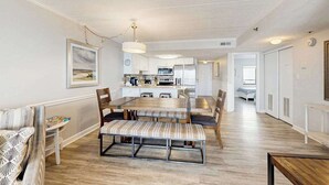 A spacious dining area with a long table and benches, a kitchen in the background, and a hallway leading to a bedroom. - Beachy Getaway Steps From Water w/Shared Pool, WiFi, AC, & Private Washer/Dryer (Ocean City)