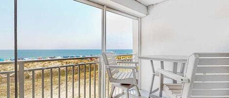 Uma varanda com vista para a praia, cadeiras brancas e uma mesa.