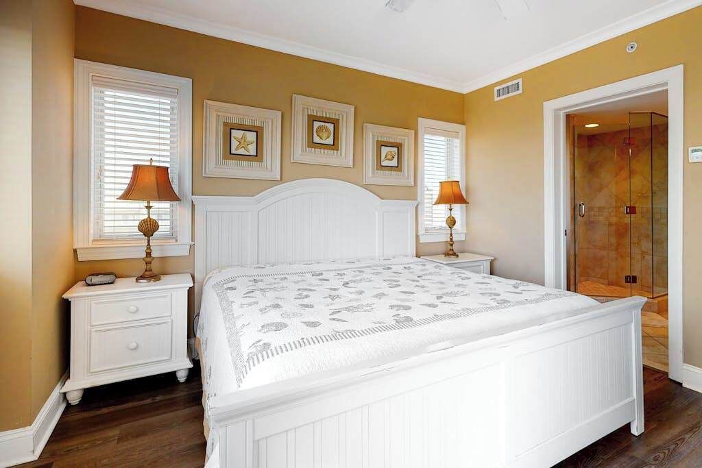 A bedroom with a white bed, two bedside tables with lamps, a large window with blinds, and a door leading to a bathroom.