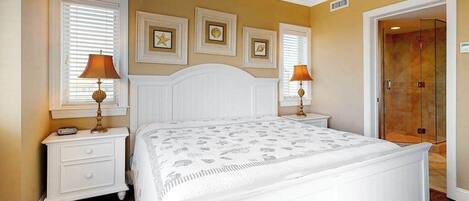 A bedroom with a white bed, two bedside tables with lamps, a large window with blinds, and a door leading to a bathroom.