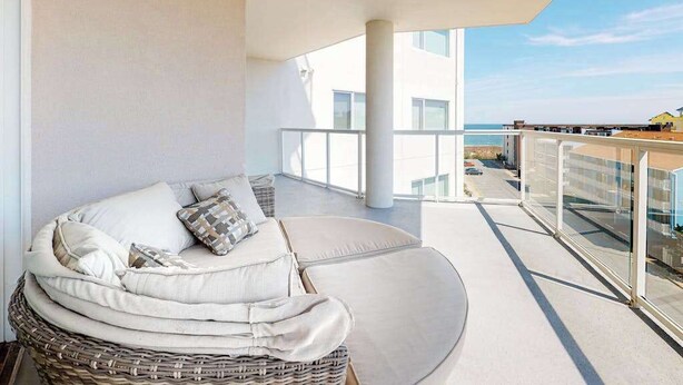 A balcony with a wicker sofa, a glass railing, and a view of the sea and buildings.