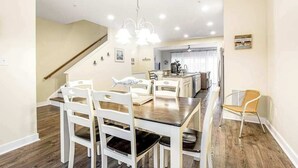A dining area with a wooden table and white chairs, a staircase, and a kitchen in the background. - Seaside Village - Bowline 13002-4 (West Ocean City)