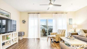 A living room with a flat-screen TV, a ceiling fan, a sofa, a coffee table, and a view through a glass door. - Seaside Village - Bowline 13002-4 (West Ocean City)
