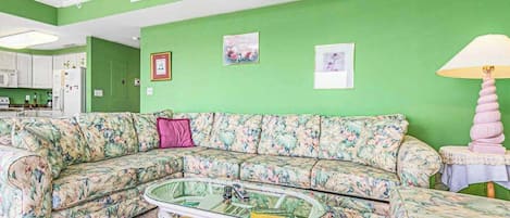 A living room with a green wall, a floral sofa, a glass coffee table, and a pink lamp.