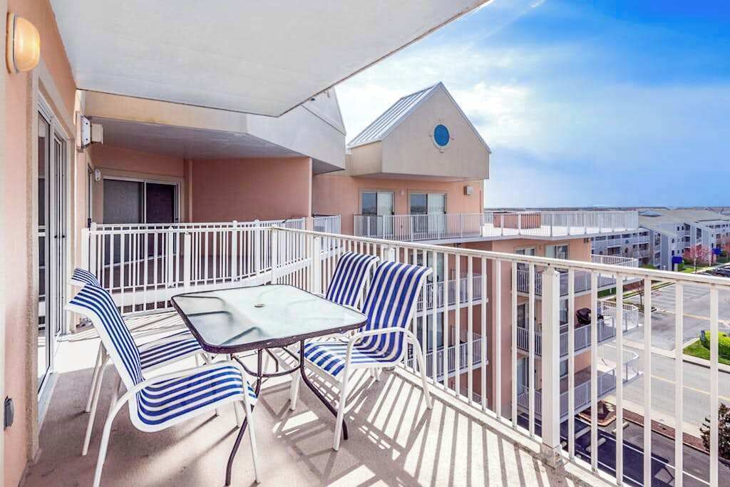 Un balcon avec une table et des chaises, une balustrade blanche et, en arrière-plan, un immeuble doté d’une fenêtre circulaire bleue.