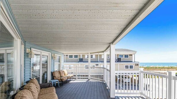 Ein überdachter Außenbalkon mit einer Couch und Stühlen, Blick aufs Meer und ein Gebäude im Hintergrund.
