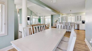 Un'ampia sala da pranzo con un lungo tavolo e sedie bianche, una cucina con mobili bianchi e una ventola a soffitto.