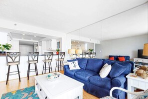 A modern living room with a blue sofa, white coffee table, and a dining area with bar stools. - Oceanfront Century I Condo w/ Ocean & Bay Views, WiFi, AC, W/D, Shared Pool/Gym (Ocean City)