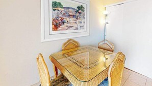 A dining area with a glass-top table and wicker chairs, a framed painting on the wall, and a wall-mounted light fixture. - Eastwinds 505 (Ocean City)