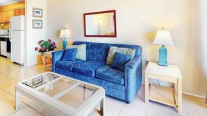 A living room with a blue sofa, a glass coffee table, a small side table with a lamp, and a kitchen in the background. - Eastwinds 505 (Ocean City)