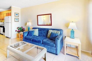 A living room with a blue sofa, a glass coffee table, a small side table with a lamp, and a kitchen in the background. - Eastwinds 505 (Ocean City)